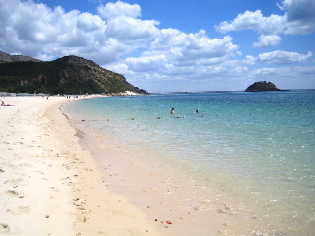 A scenic beach with clear turquoise waters and people swimming, with a small island in the distance and a mountain to the left, under a blue sky dotted with fluffy clouds.