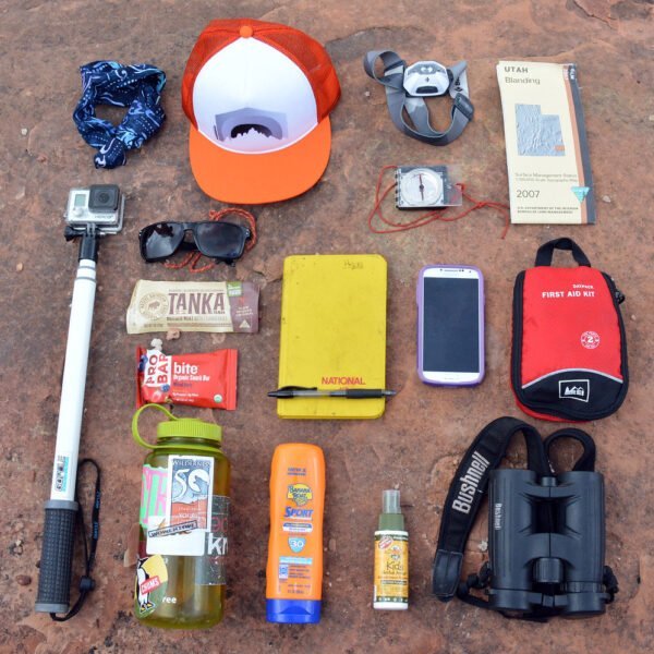 Various hiking essentials laid out on a rock surface, including a bandana, trucker hat, sunglasses, hiking map, camera on a selfie stick, energy bars, a yellow notebook, two smartphones, a first aid kit, water bottle with stickers, sunscreen, insect repellent spray, and a pair of binoculars.