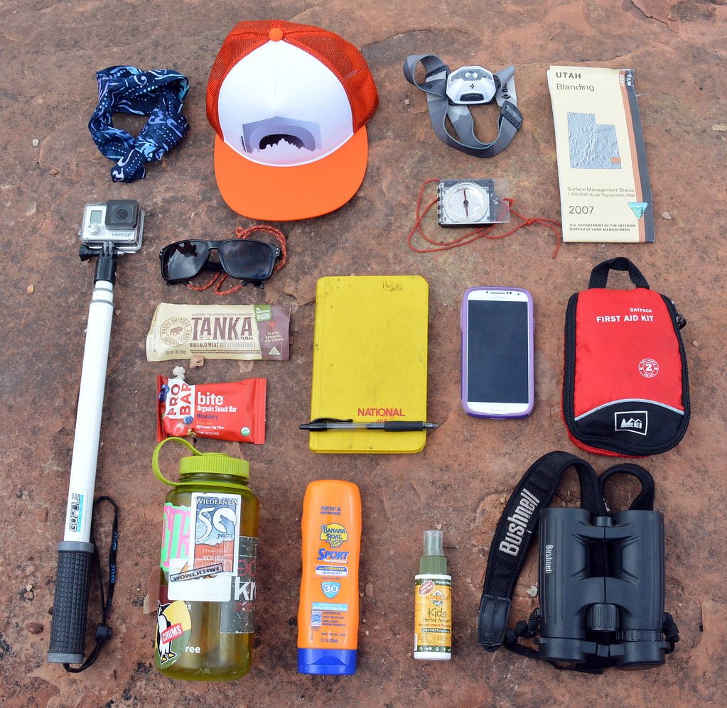 Various hiking essentials laid out on a rock surface, including a bandana, trucker hat, sunglasses, hiking map, camera on a selfie stick, energy bars, a yellow notebook, two smartphones, a first aid kit, water bottle with stickers, sunscreen, insect repellent spray, and a pair of binoculars.