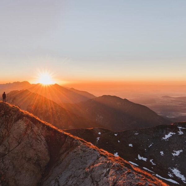 A person standing on a mountain ridge at sunrise with the sun casting a warm glow over the surrounding peaks and valleys.