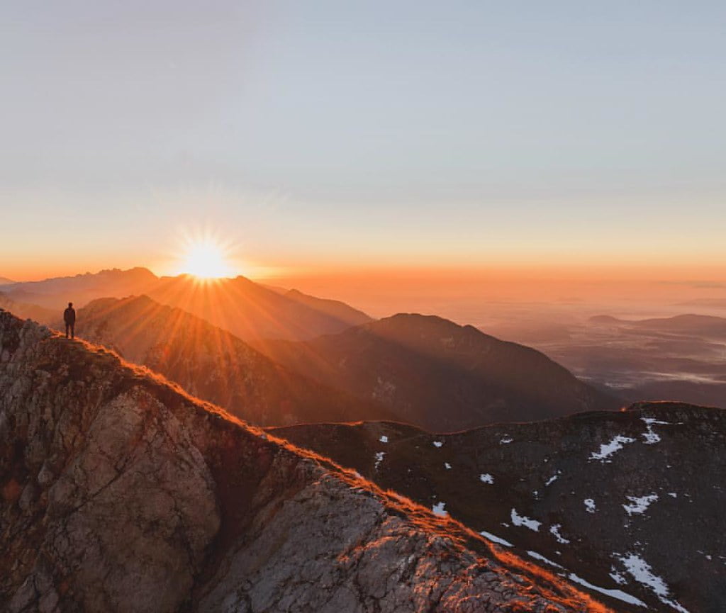 A person standing on a mountain ridge at sunrise with the sun casting a warm glow over the surrounding peaks and valleys.