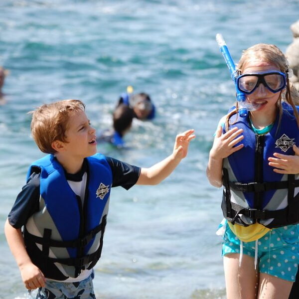 A boy and a girl in life jackets standing by the sea, with the girl wearing snorkeling gear. People are swimming in the background.