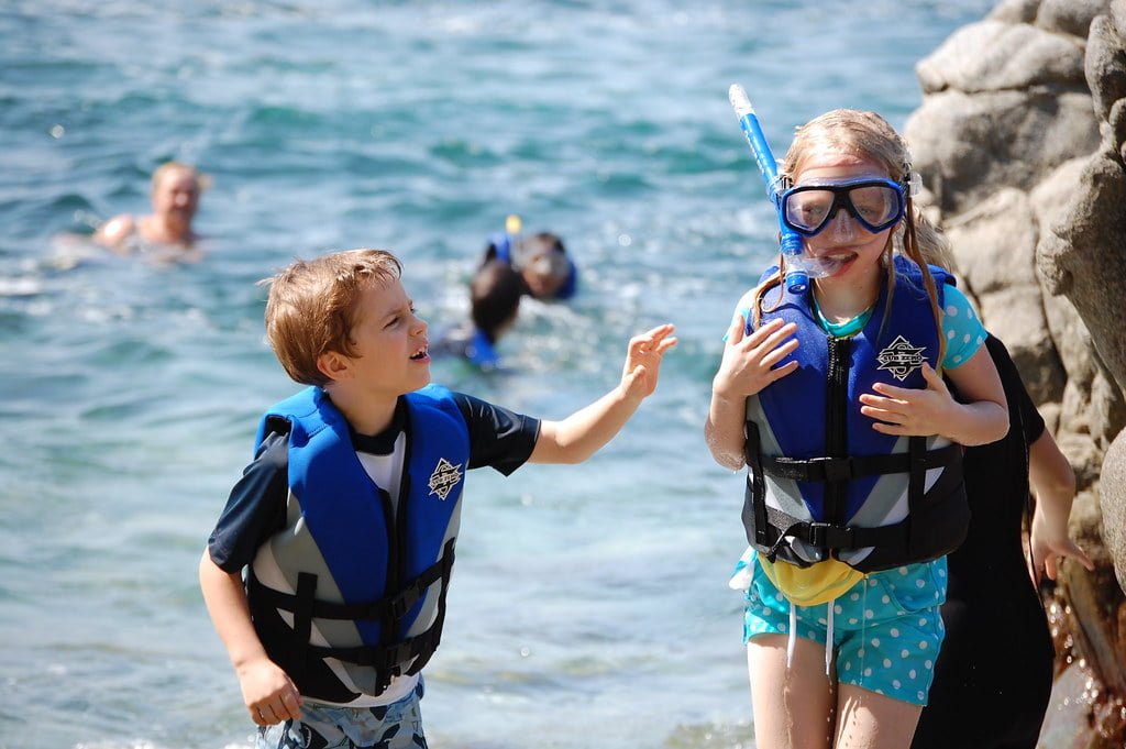 A boy and a girl in life jackets standing by the sea, with the girl wearing snorkeling gear. People are swimming in the background.