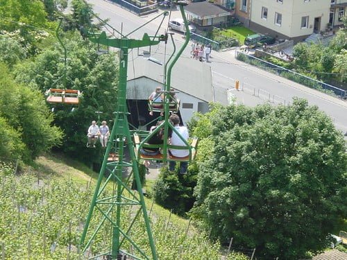 Chairlift with passengers over green trees with a road and buildings in the background.