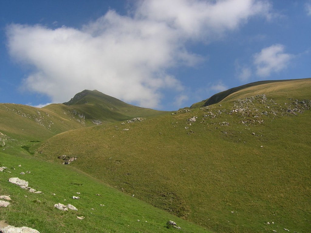 A lush green hillside with a clear blue sky and scattered clouds, with a ridge leading up to a peak in the distance.
