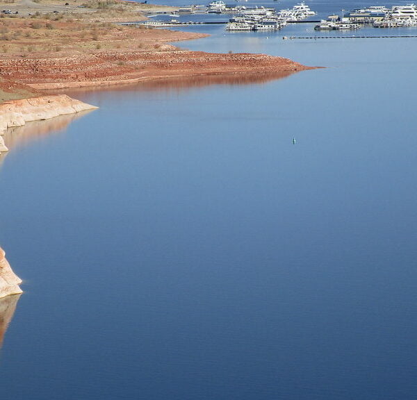 A calm blue reservoir with a visible waterline on the surrounding red cliffs, accompanied by a marina with multiple boats docked on the right side.