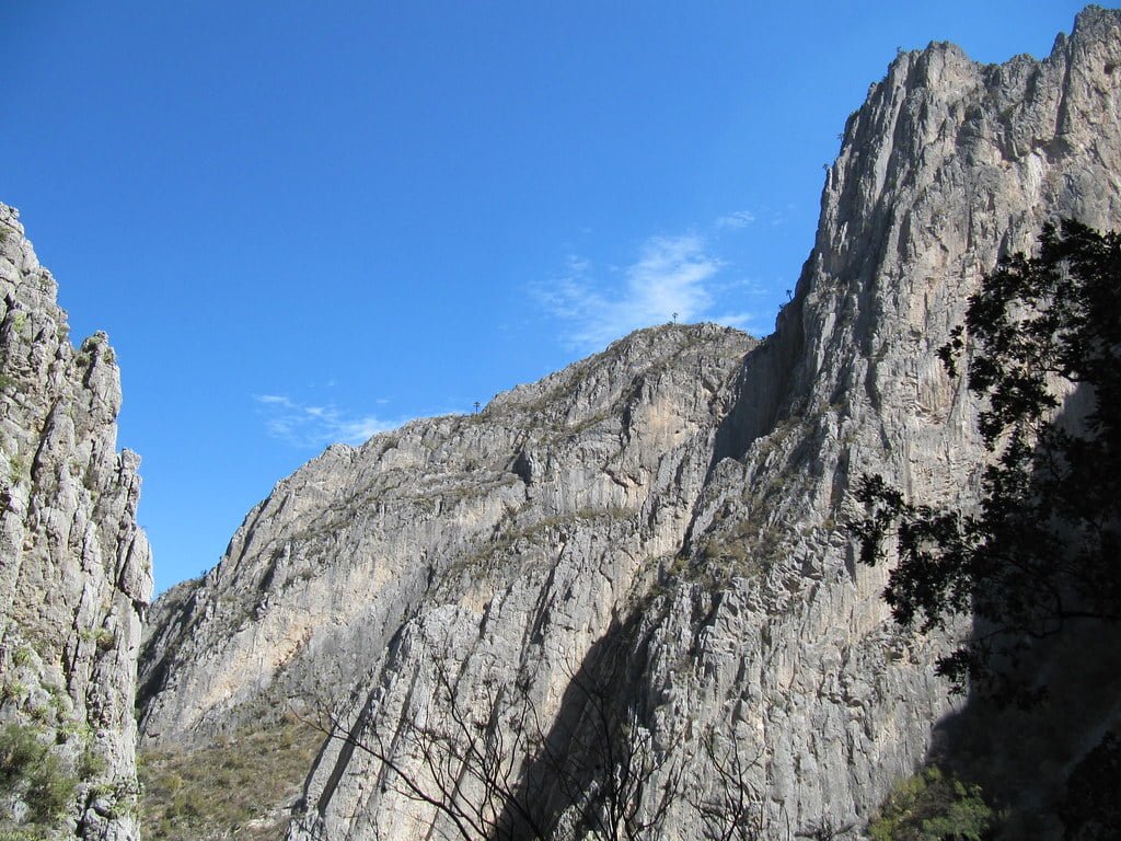 Steep, rugged mountain cliffs under a clear blue sky, with sparse vegetation and shadows from surrounding trees.
