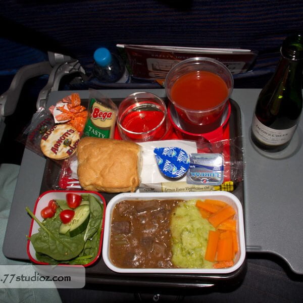 Airplane meal setup on a tray table with a laptop to the side; includes a salad, a bread roll, butter, cheese, main course with beef stew, mashed potatoes and carrots, crackers, dried apricots, a cup of water, a cup of orange juice, red jelly dessert, and a small bottle of wine.