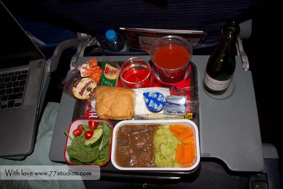 Airplane meal setup on a tray table with a laptop to the side; includes a salad, a bread roll, butter, cheese, main course with beef stew, mashed potatoes and carrots, crackers, dried apricots, a cup of water, a cup of orange juice, red jelly dessert, and a small bottle of wine.