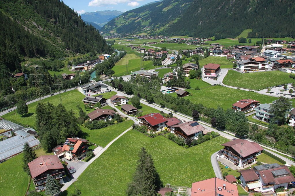 Aerial view of a lush green alpine valley with a river running through it, featuring scattered houses with red roofs, a church spire, and surrounding forested mountains.