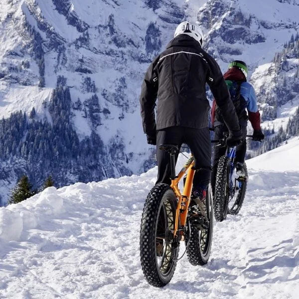 Two people mountain biking on a snow-covered trail with a backdrop of snow-capped mountains and pine trees.