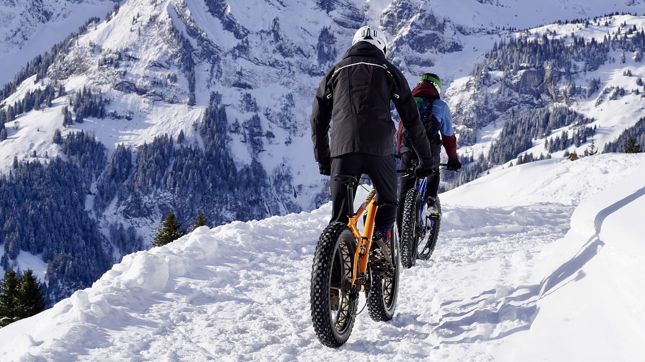 Two people mountain biking on a snow-covered trail with a backdrop of snow-capped mountains and pine trees.