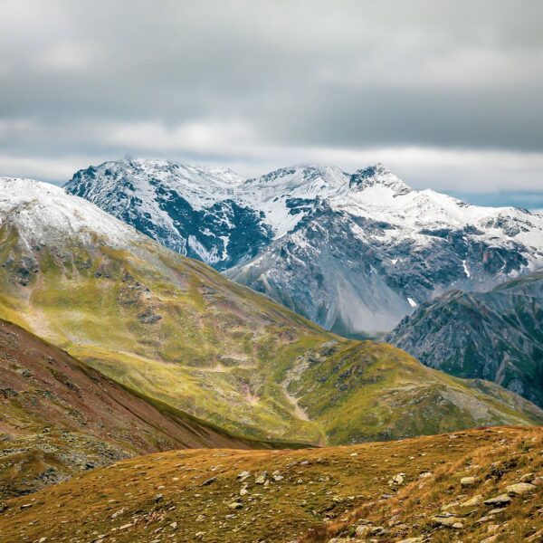 A sweeping view of rugged mountains with snow-capped peaks under a cloudy sky, foreground showing grassy slopes with patches of snow.