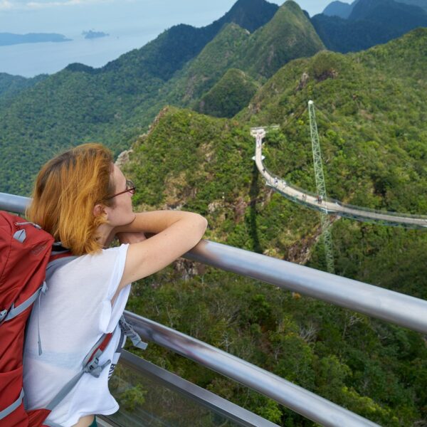 A traveler with a red backpack leans on a railing, looking out at a scenic view of a sky bridge winding through lush green mountains and the ocean in the distance.