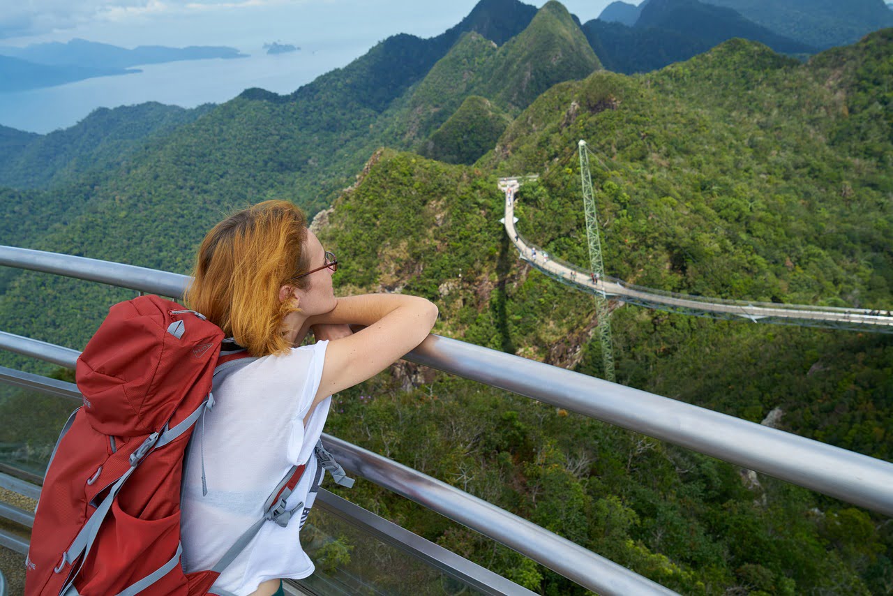 A traveler with a red backpack leans on a railing, looking out at a scenic view of a sky bridge winding through lush green mountains and the ocean in the distance.