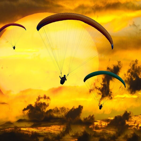 Three paragliders flying against a dramatic sunset with a backdrop of vibrant orange clouds and a large sun partially obscured by clouds.