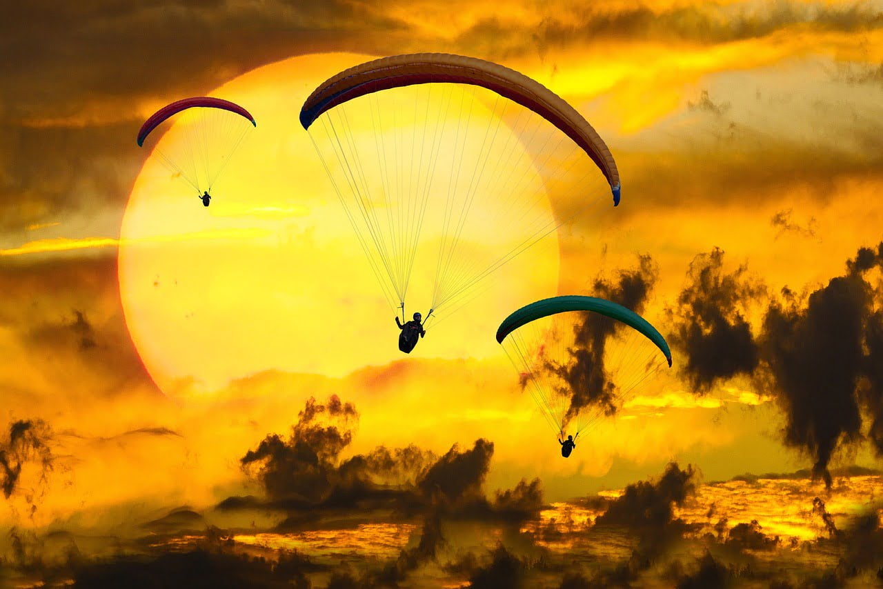 Three paragliders flying against a dramatic sunset with a backdrop of vibrant orange clouds and a large sun partially obscured by clouds.