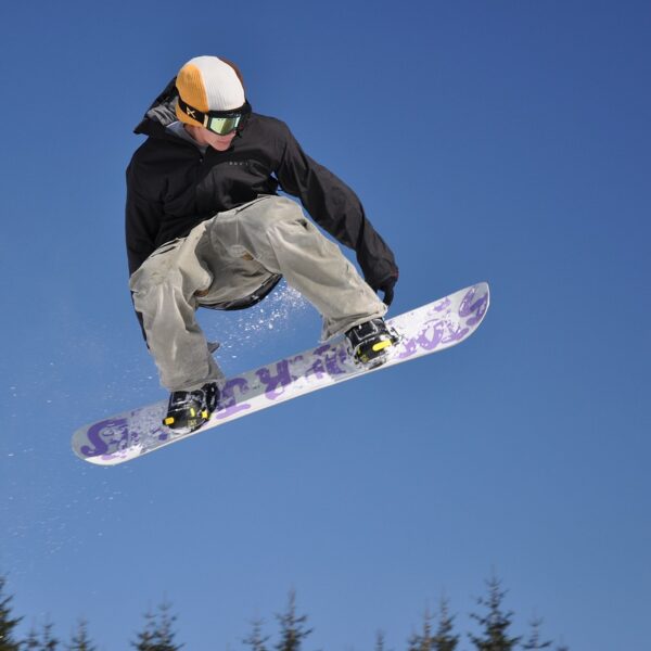 A snowboarder performing a jump against a clear blue sky with snow-covered trees in the background.