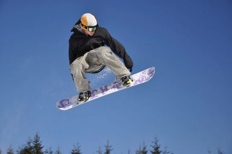A snowboarder performing a jump against a clear blue sky with snow-covered trees in the background.