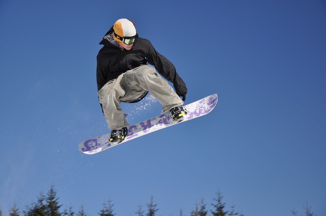 A snowboarder performing a jump against a clear blue sky with snow-covered trees in the background.