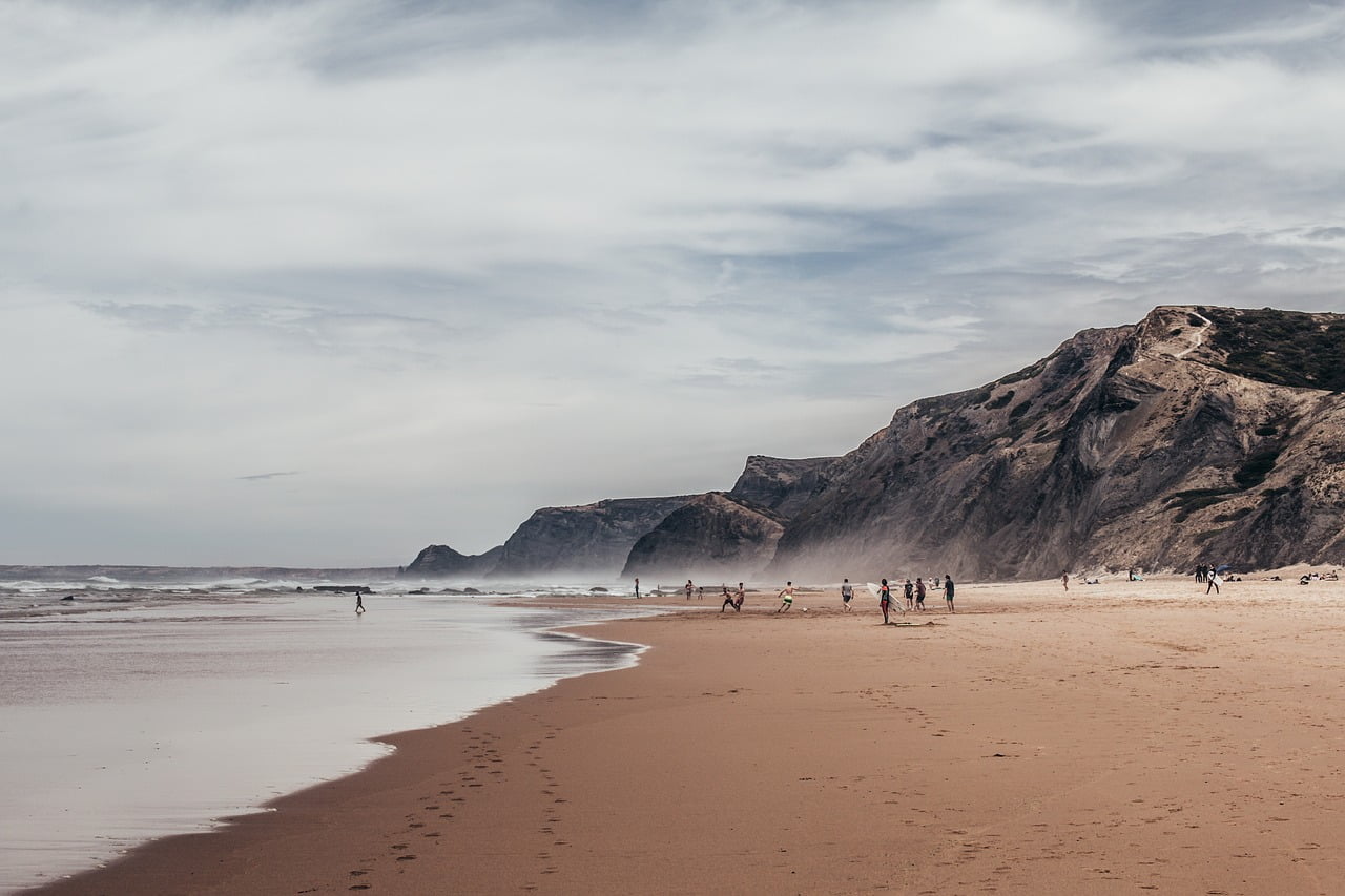 A sandy beach with people walking and playing near the water, with a backdrop of large cliffs and a hazy sky.