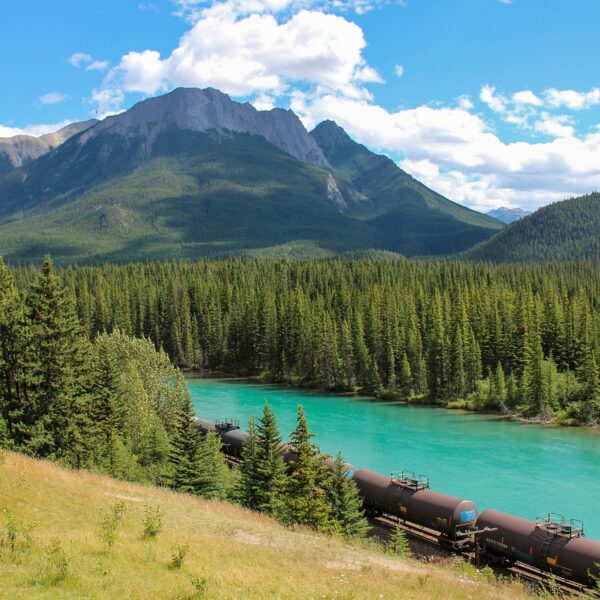 A train of black tank cars running alongside a vibrant turquoise river, with dense evergreen forests and a majestic mountain backdrop under a partly cloudy sky.