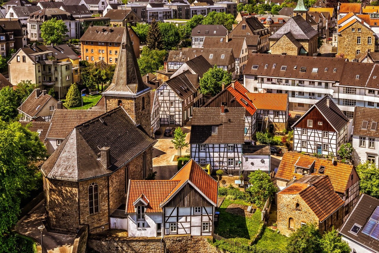 Aerial view of a quaint European village showcasing a mix of traditional half-timbered houses and stone buildings with steeply pitched roofs, alongside modern structures, narrow streets, greenery, and a church spire.