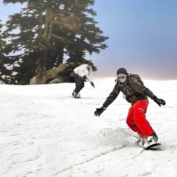 Two snowboarders descending a snow-covered slope with evergreen trees in the background and a hazy sky above. The foreground snowboarder is in focus, wearing a black jacket and red pants, carving a turn with snow spraying to the side.