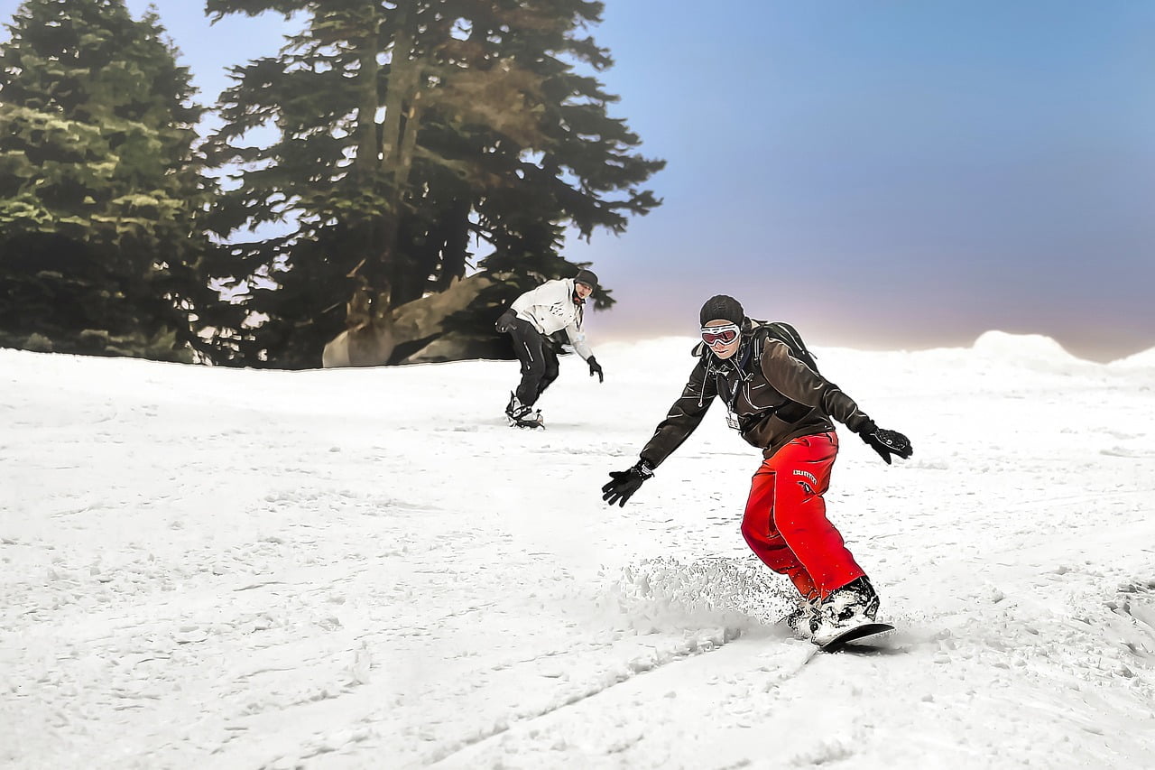 Two snowboarders descending a snow-covered slope with evergreen trees in the background and a hazy sky above. The foreground snowboarder is in focus, wearing a black jacket and red pants, carving a turn with snow spraying to the side.