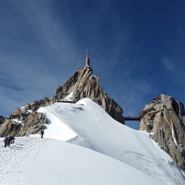 A group of climbers ascending a steep snow-covered slope towards a rocky mountain peak with a structure and antenna on top, under a clear blue sky.