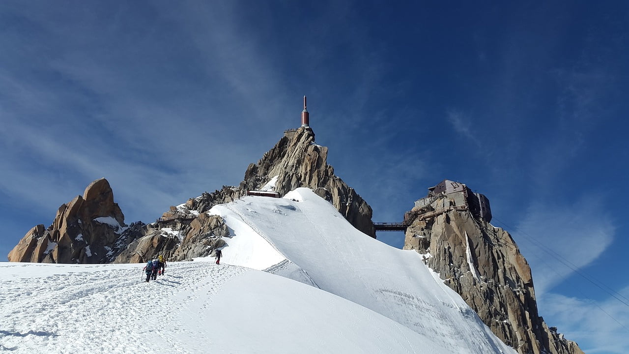 A group of climbers ascending a steep snow-covered slope towards a rocky mountain peak with a structure and antenna on top, under a clear blue sky.