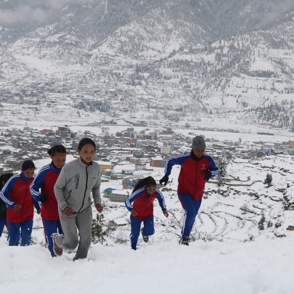 A group of children in sportswear walking through snow with a mountainous village in the background.