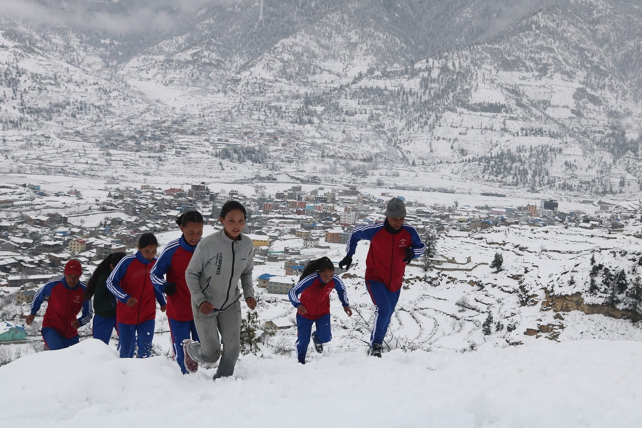 A group of children in sportswear walking through snow with a mountainous village in the background.