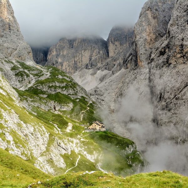 A small mountain refuge sits nestled on a green slope in a dramatic alpine landscape with steep rocky cliffs and swirling mist.