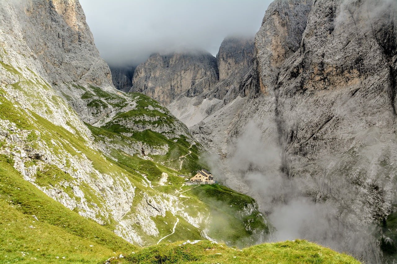 A small mountain refuge sits nestled on a green slope in a dramatic alpine landscape with steep rocky cliffs and swirling mist.