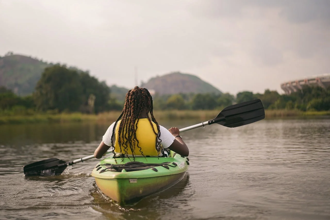 A person with braided hair wearing a yellow life jacket is kayaking on a calm river, with lush greenery and hills in the background.