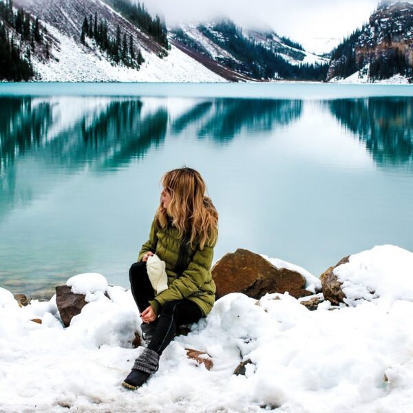 A person sitting on snow-covered rocks beside a tranquil turquoise lake with snow-dusted mountains reflected in the water.