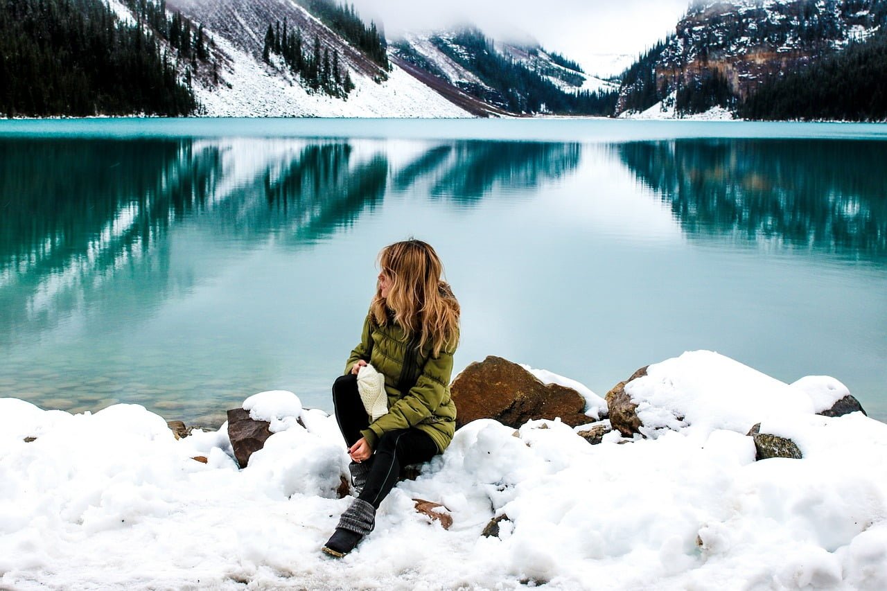 A person sitting on snow-covered rocks beside a tranquil turquoise lake with snow-dusted mountains reflected in the water.