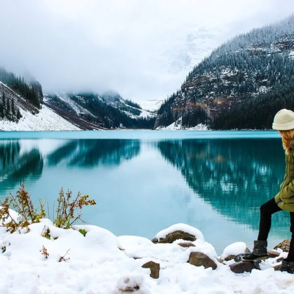 A person in a green coat and white hat sitting on a rock wall next to a serene turquoise lake with snowy mountainous background and fog.