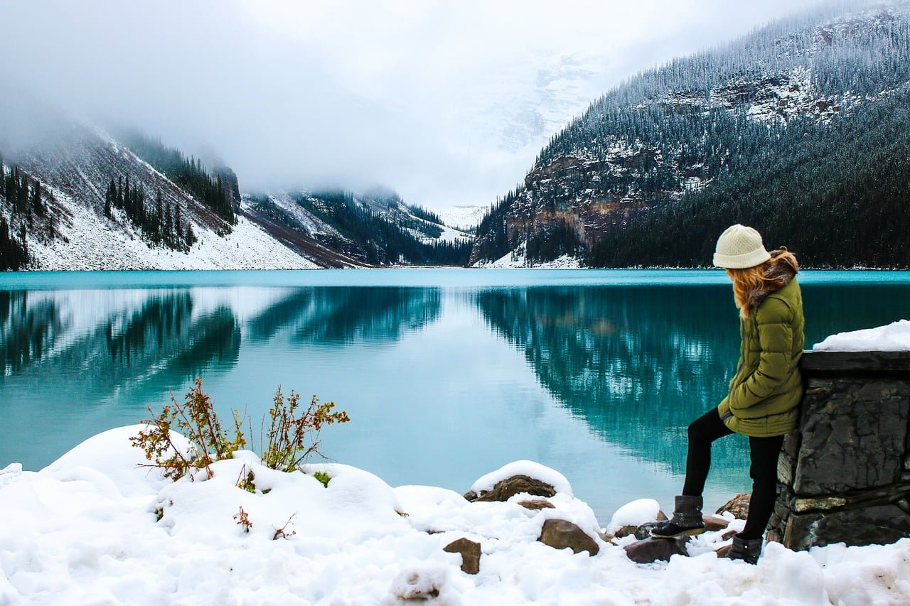 A person in a green coat and white hat sitting on a rock wall next to a serene turquoise lake with snowy mountainous background and fog.