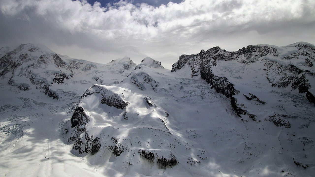 A panoramic view of a snow-covered mountain range with peaks and ridges under a cloudy sky.