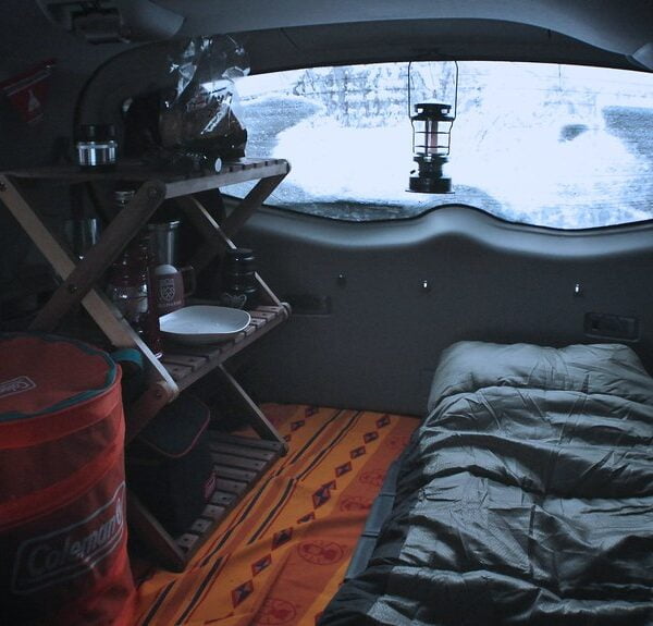 Interior of a camper van with a sleeping bag on a bed, a wooden table with a stove, a cooler, and a lantern, viewed from the inside with a snowy landscape visible through the windshield.