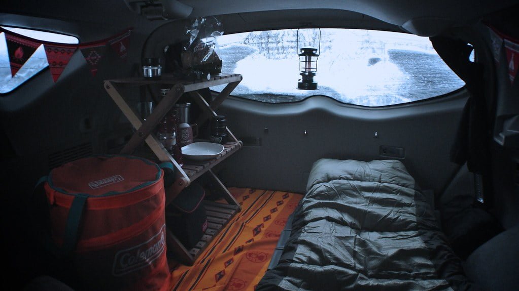 Interior of a camper van with a sleeping bag on a bed, a wooden table with a stove, a cooler, and a lantern, viewed from the inside with a snowy landscape visible through the windshield.