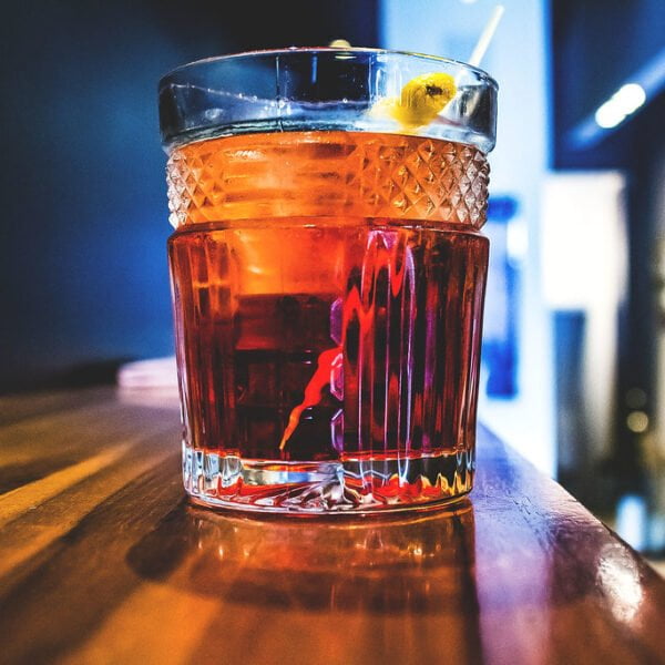 A close-up of a whiskey cocktail with ice and a lemon twist, in a textured glass, placed on a wooden bar with a blurred background.