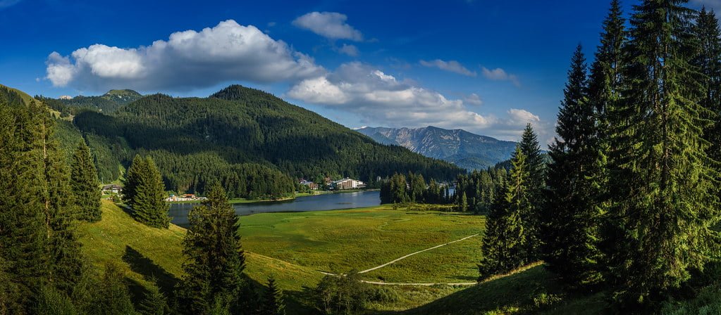 Panoramic view of a mountain lake surrounded by evergreen forests with a clear sky and fluffy clouds above.