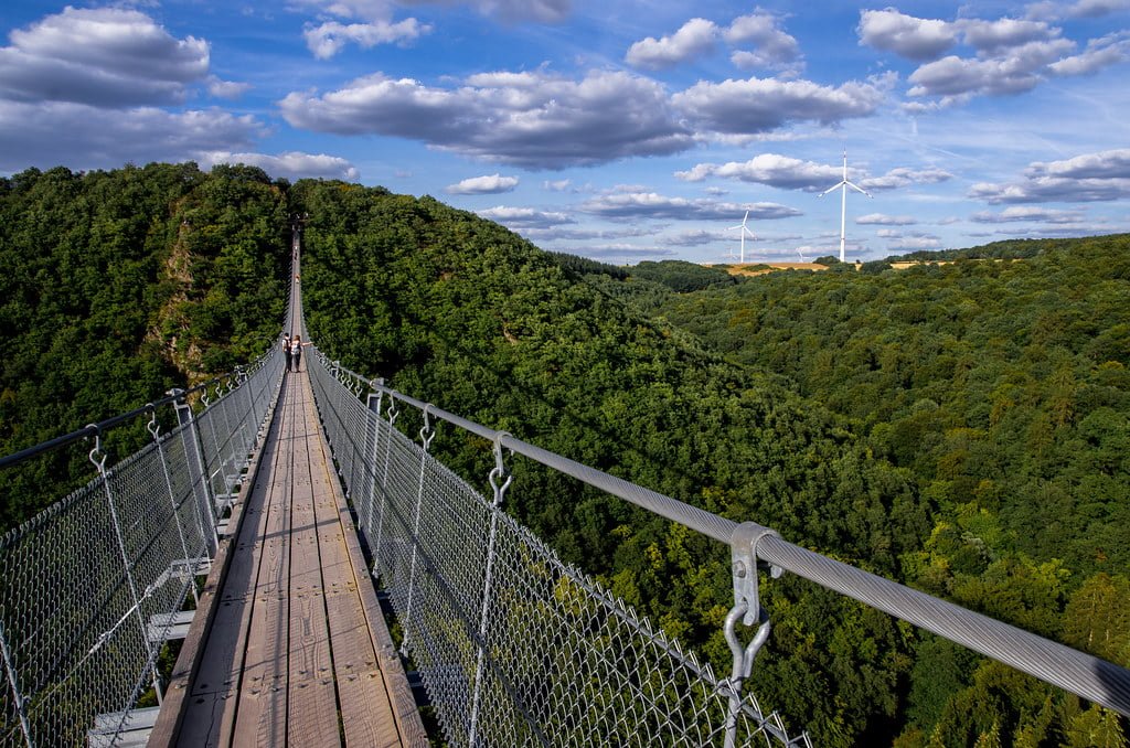 A suspension bridge with a wooden walkway spans across a lush green valley, with visitors walking on it and wind turbines visible on the horizon under a partly cloudy sky.