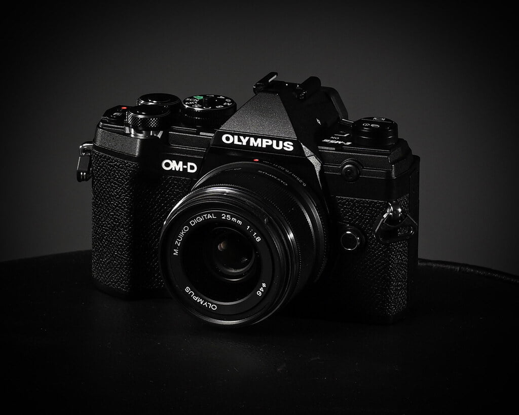 An Olympus OM-D series camera with a M.Zuiko Digital 25mm 1:1.8 lens positioned against a black background.