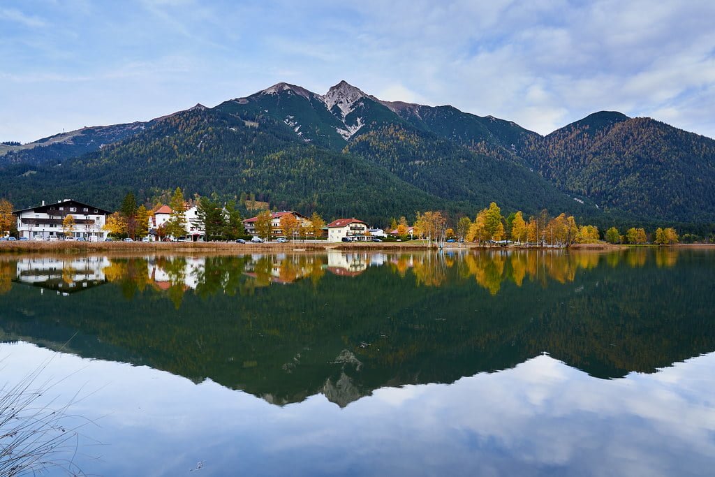 A calm lake reflecting a small village with autumn-colored trees and a mountain backdrop under a blue sky with wispy clouds.