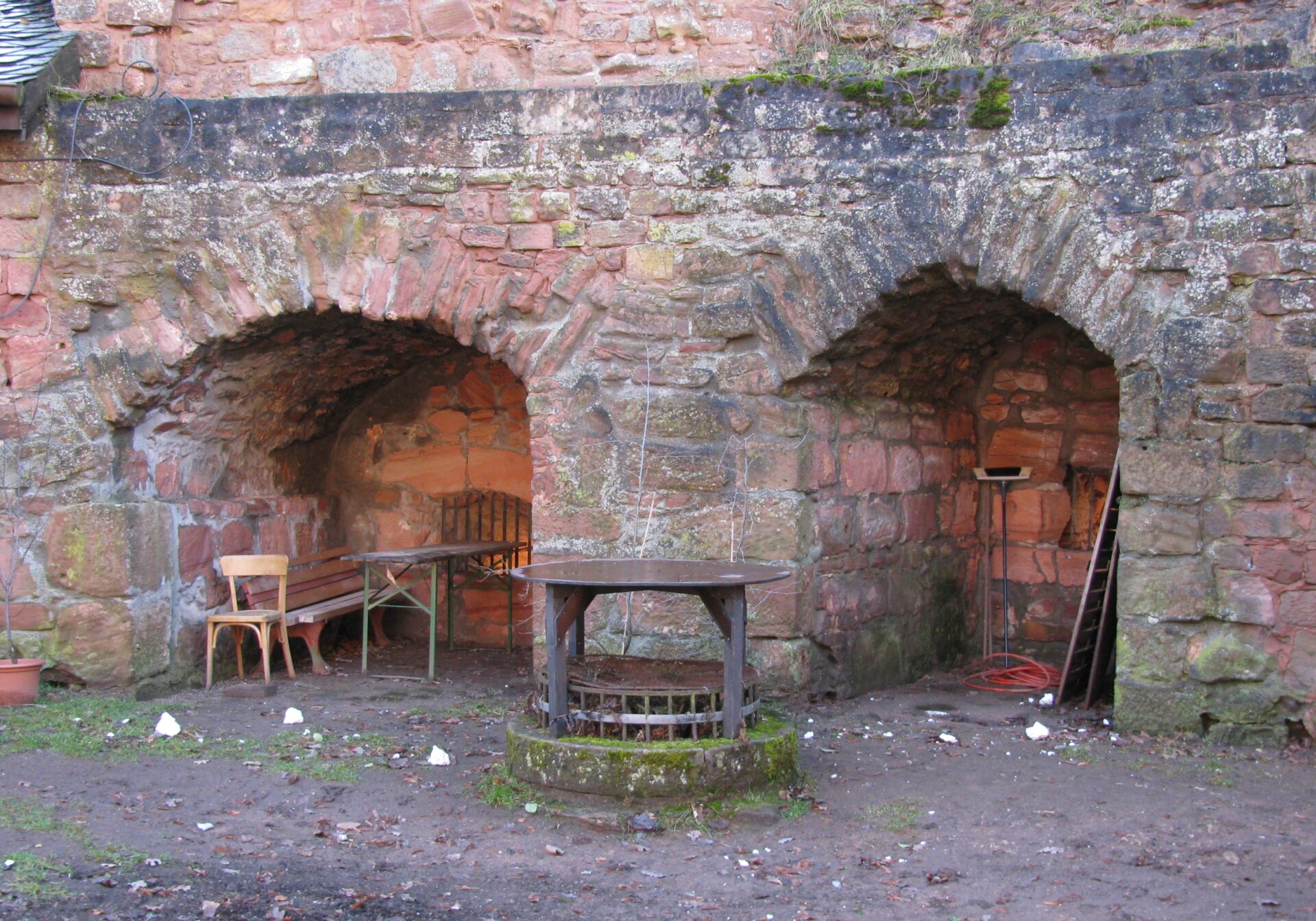 Two arched alcoves in an old red brick wall, with an empty table and chairs on the left and a round well-like structure in the center. There is scattered debris on the ground.