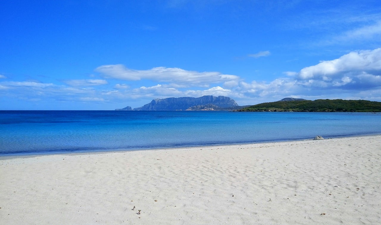 A tranquil beach scene with white sands in the foreground, clear blue water in the middle, a mountain range on the horizon, and a sky with scattered clouds above.